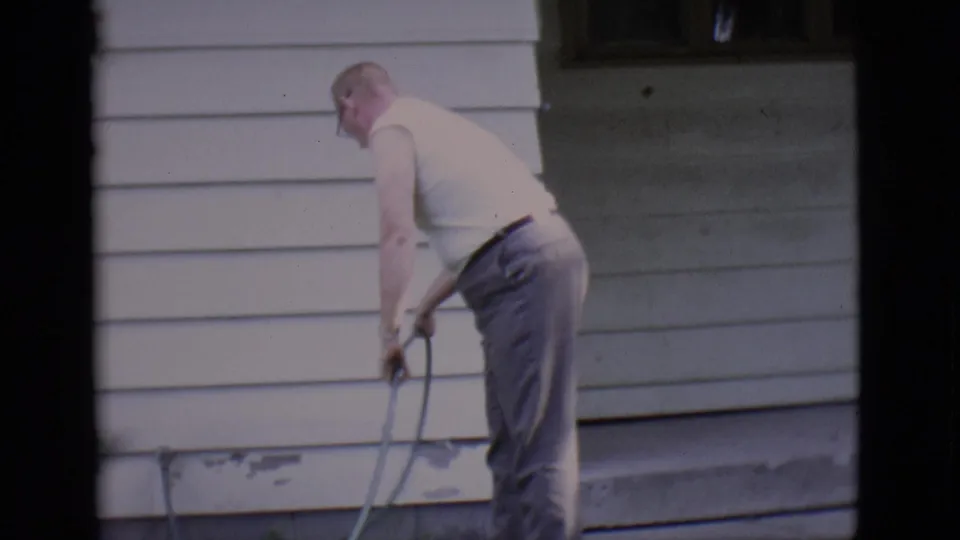 An adult man in a white sleeveless shirt manages a garden hose against the siding of a Clarkdale house in 1966 footage.