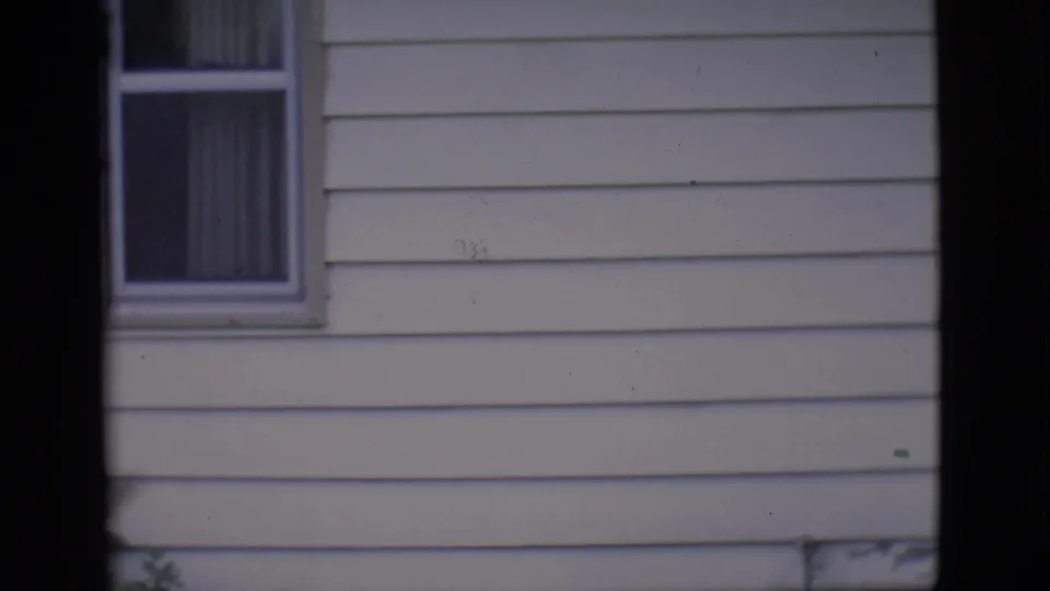 The final frame holds on the white-sided house exterior and curtained window, with the people now out of view.