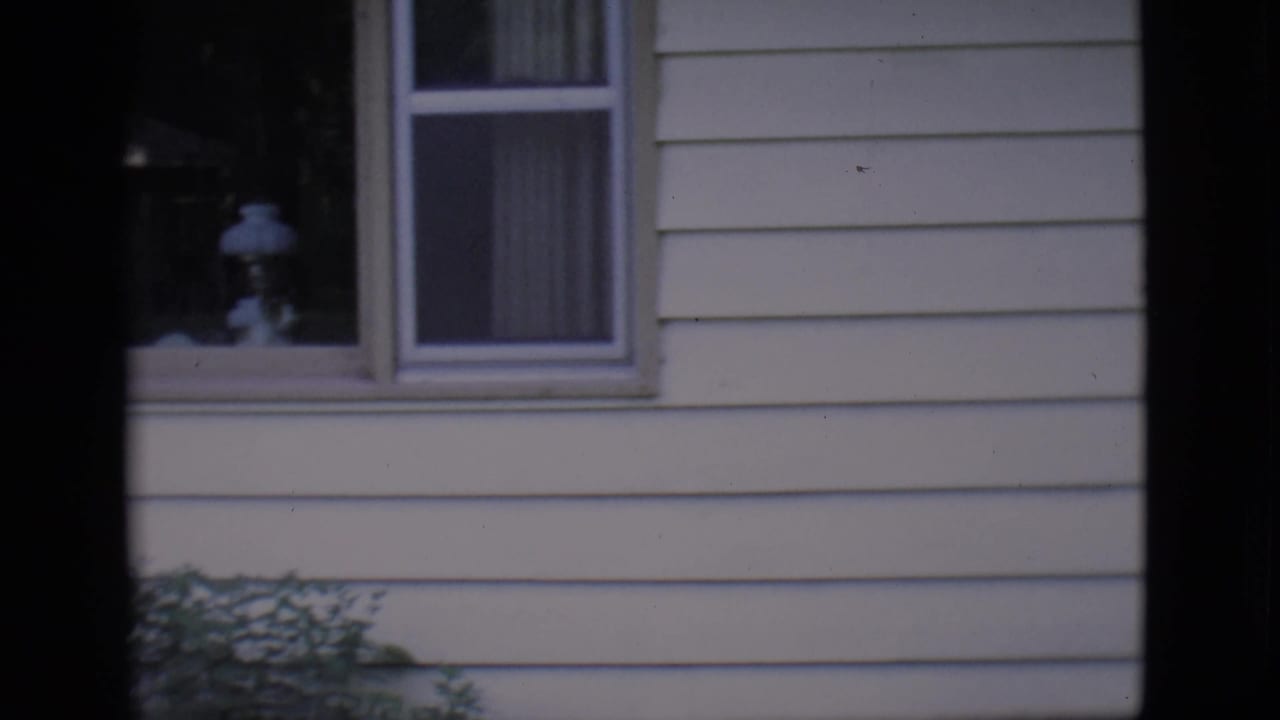 Light-colored house exterior with windows, curtains, shrubs, and dark film-frame borders.