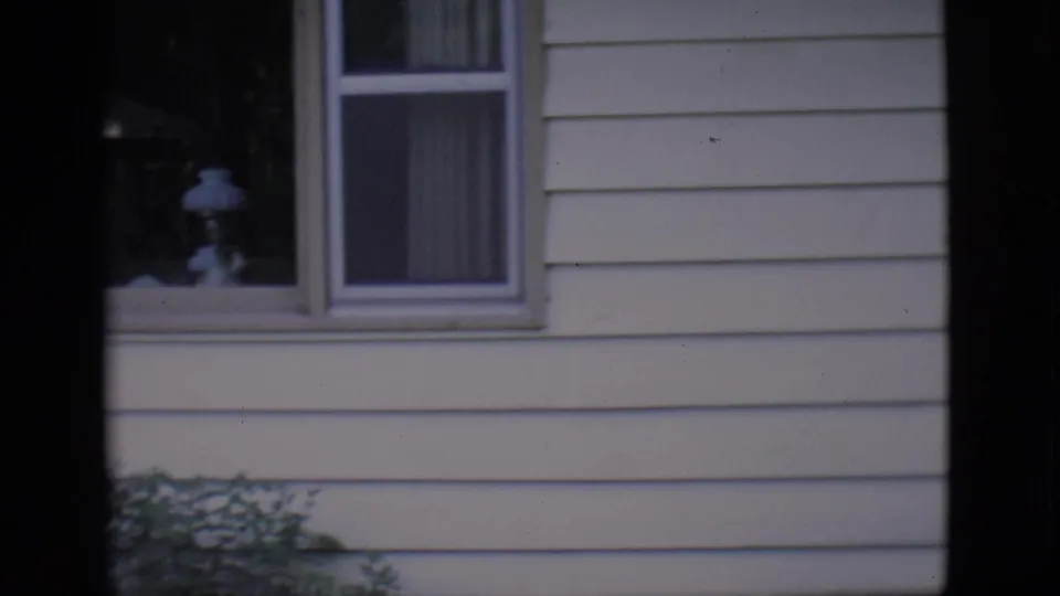 1966 Clarkdale home-movie frame showing a light-sided house exterior with curtained windows, shrubs, and visible film-frame borders.