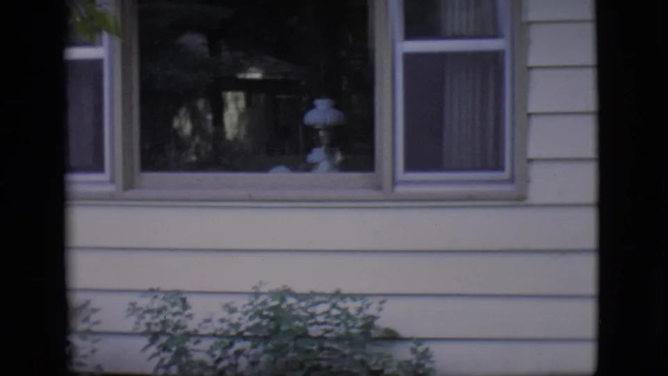 1966 Clarkdale frame of a residential exterior with horizontal siding, curtained windows, an interior lamp, shrubs, and film borders.