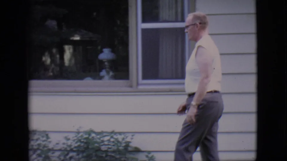 1966 Clarkdale home-movie frame showing an adult man in glasses walking left past a light-sided residential exterior.