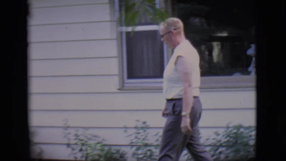 1966 Clarkdale frame with an adult man in glasses walking beside a light-colored house exterior and low shrubs.