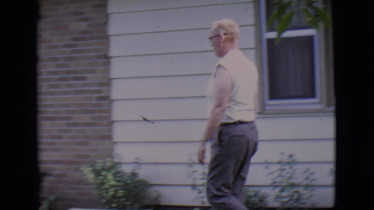 Adult man in a sleeveless shirt walks past a house exterior and shrubs.
