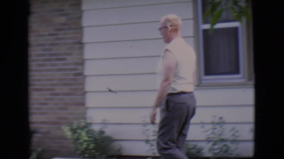 Adult man walks across a front lawn beside a brick and siding house in 1966 Clarkdale, Arizona home-movie footage.
