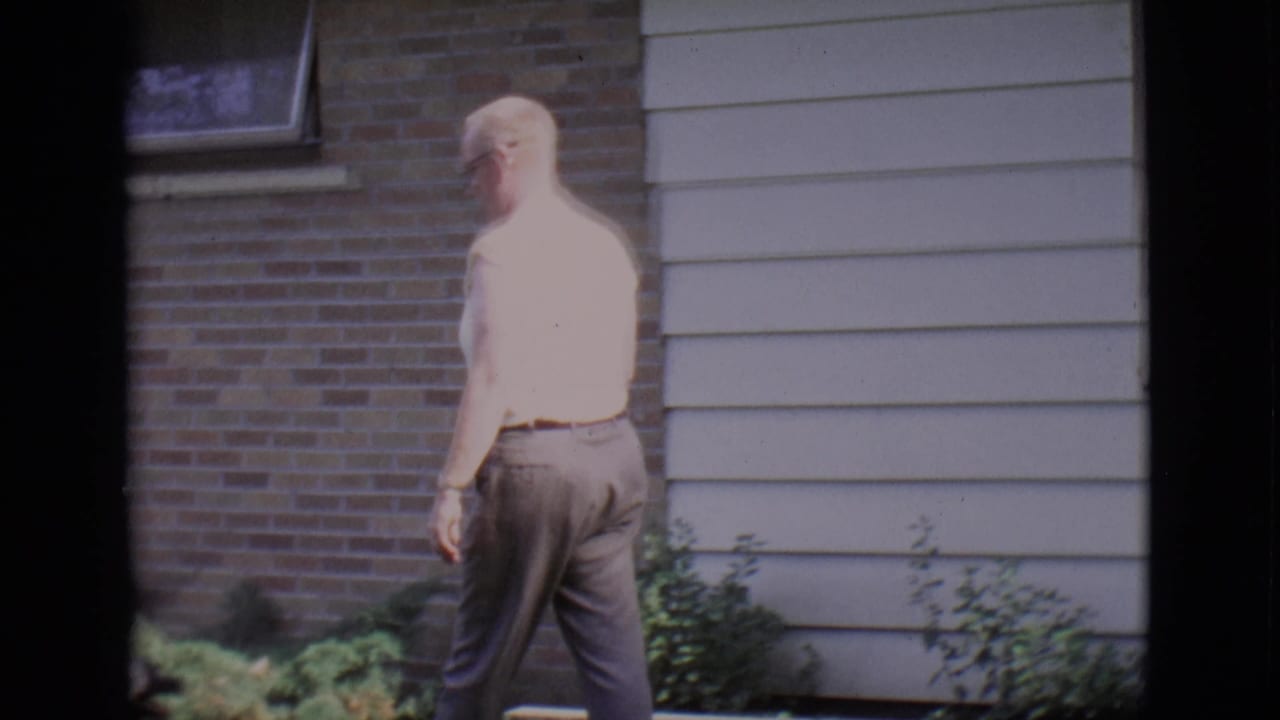 Man continues walking near a brick wall, siding, window, and low greenery.