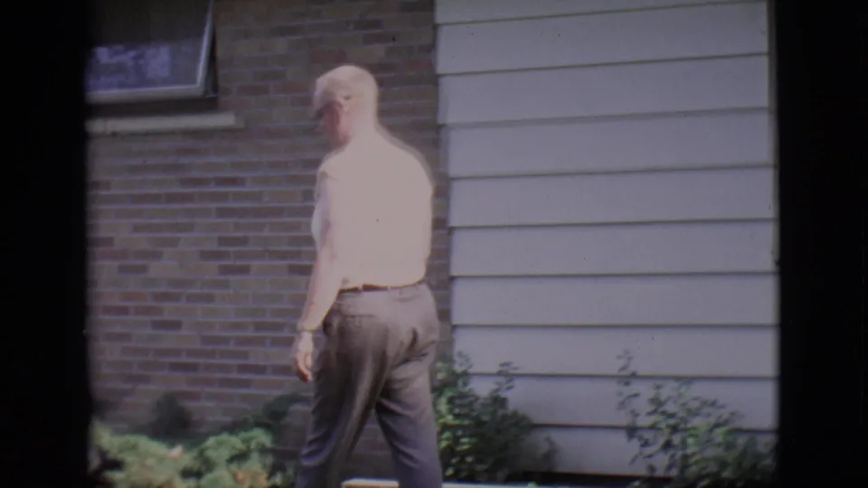 The man moves along the front lawn near a brick-and-siding house in bright 1966 Clarkdale sunlight.