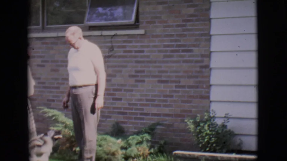 The man pauses beside the lawn and shrubbery as the dog remains partly visible in 1966 Clarkdale footage.
