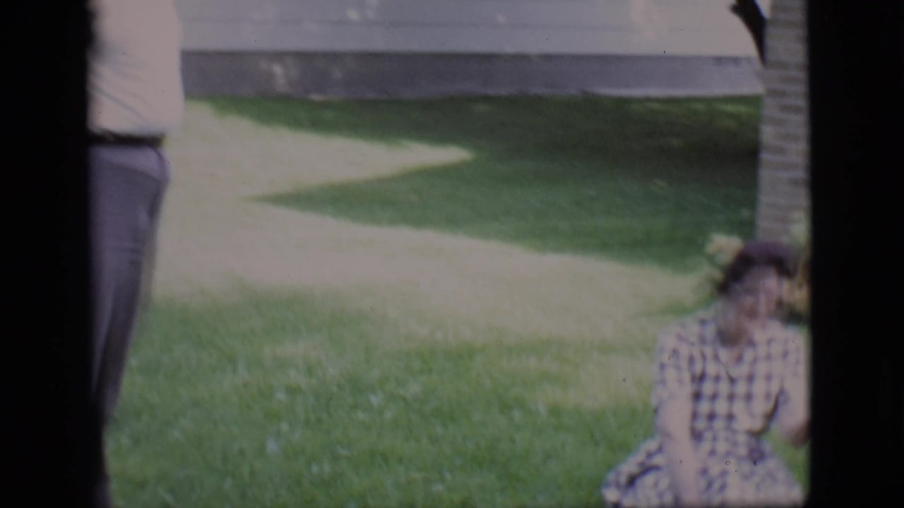 Seated woman on grass near a sidewalk, with a cropped standing figure at the frame edge.