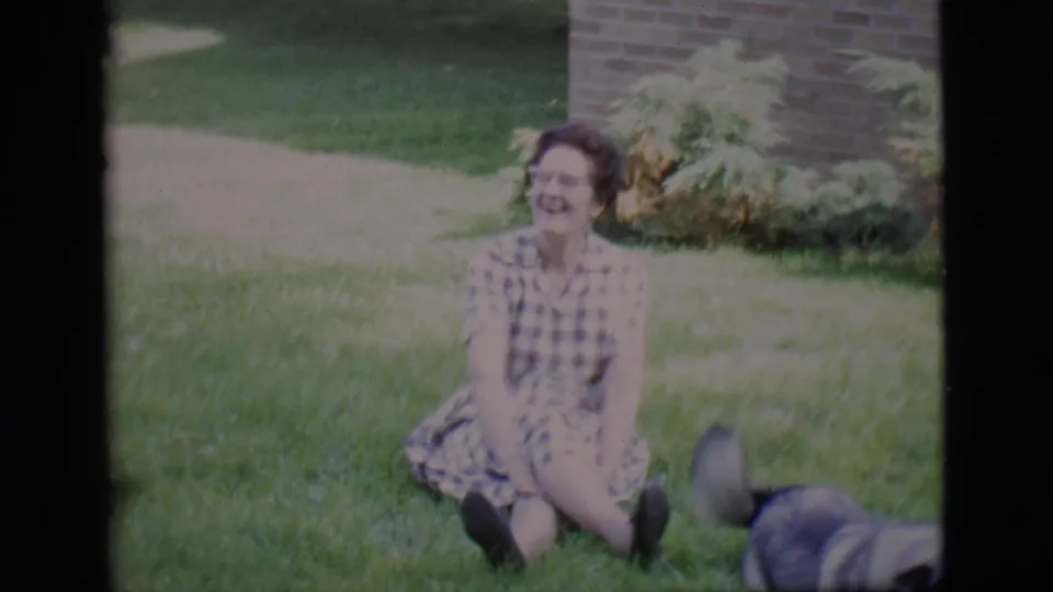 A woman in a checkered dress smiles on a Clarkdale lawn, with a dog partly visible beside her in 1966 home-movie footage.