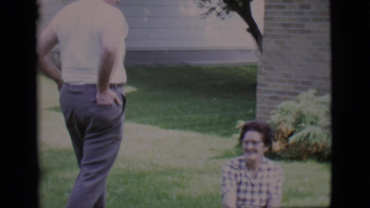 Man standing near a seated woman on a lawn beside a brick house.