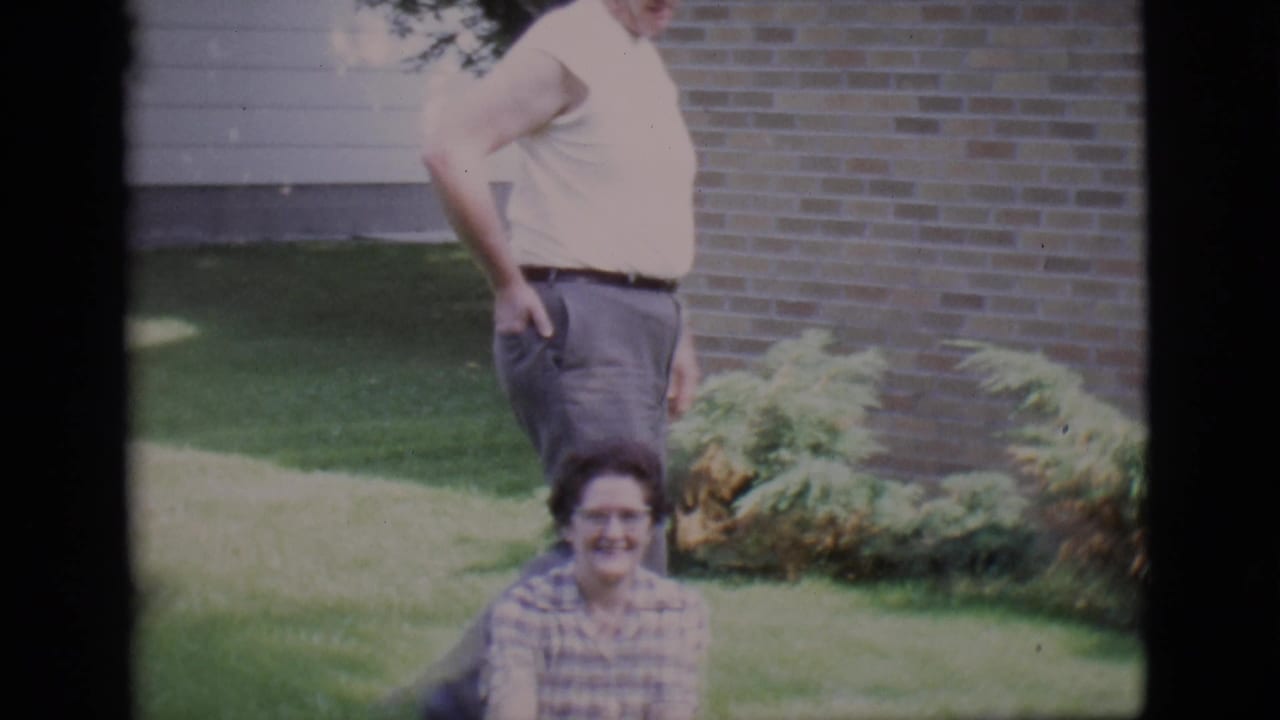 Man and seated woman in a grassy yard in front of a brick wall.