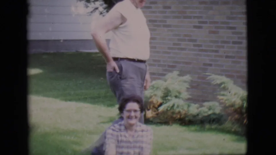 1966 Clarkdale backyard frame with a man standing over a seated woman on a lawn near shrubs and a brick house.
