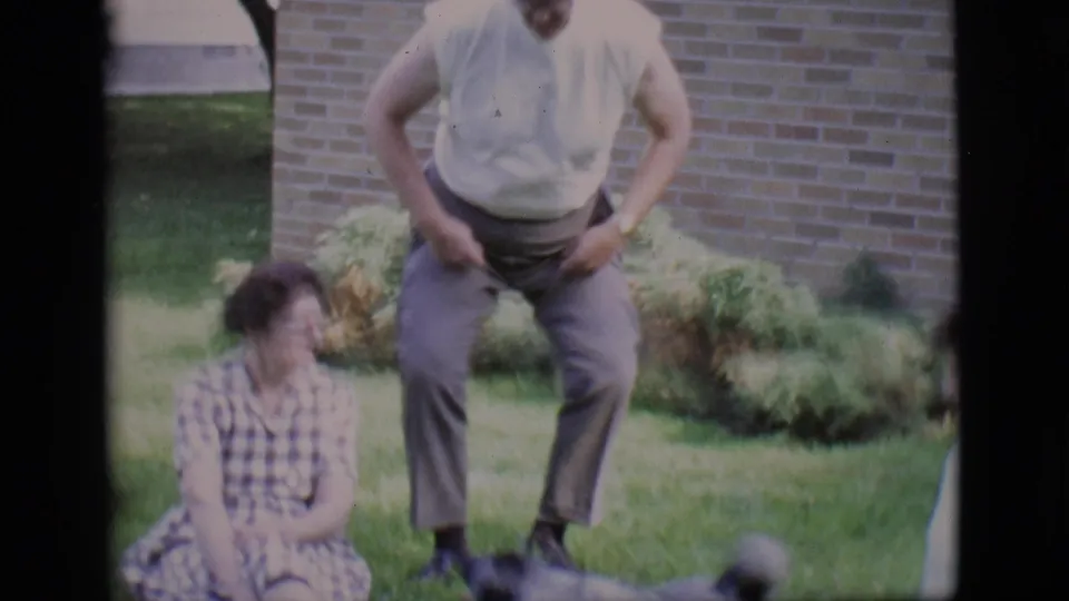 Home-movie frame from 1966 Clarkdale showing the woman seated while the man lowers himself near her in the backyard.
