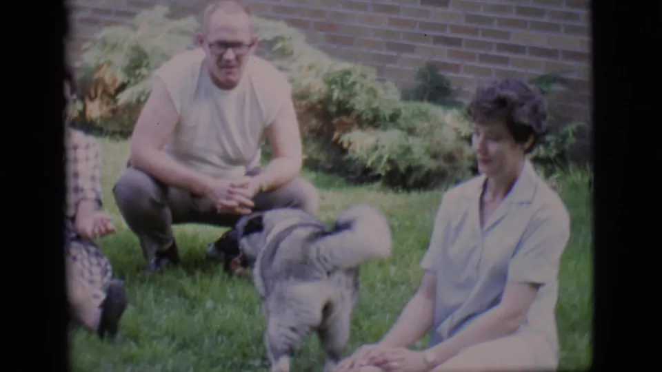 The German Shepherd crosses the lawn in front of seated adults in 1966 Clarkdale backyard footage.