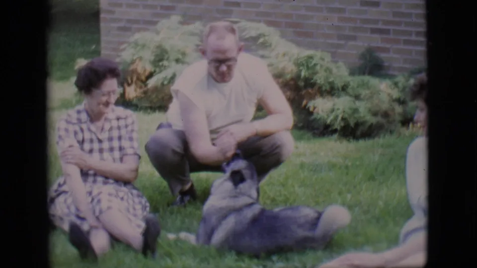 A 1966 home-movie frame from Clarkdale shows two adults on a lawn as the dog lowers itself in front of the crouching man.