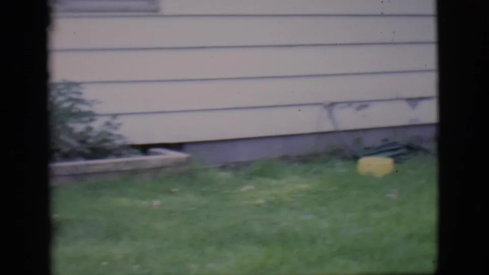 1966 Clarkdale home-movie frame showing a white-sided house exterior, green lawn, low stone border, and soft 8mm color.