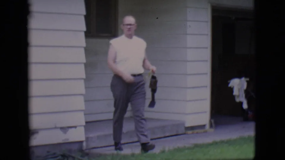 1966 Clarkdale frame of an adult man in a sleeveless white shirt and dark trousers walking from a house doorway with a belt in hand.