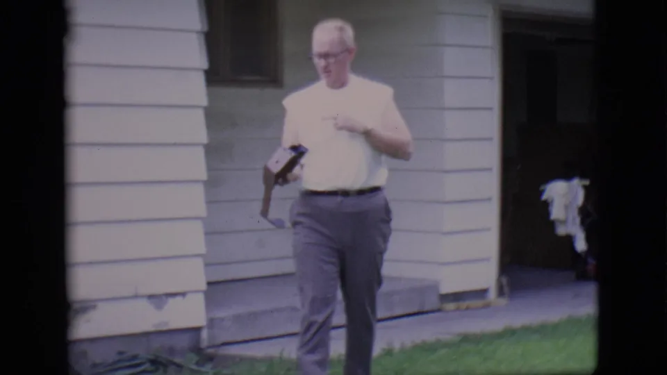 Adult man walks toward the camera outside a white-sided Clarkdale house in 1966, holding a belt as 8mm film edges frame the scene.