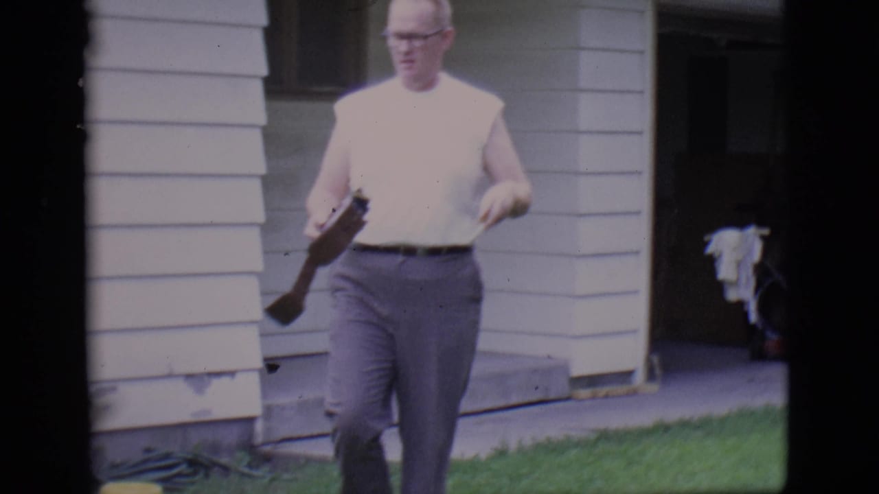 Man in a sleeveless white shirt walks on a lawn holding a small movie camera near a house.
