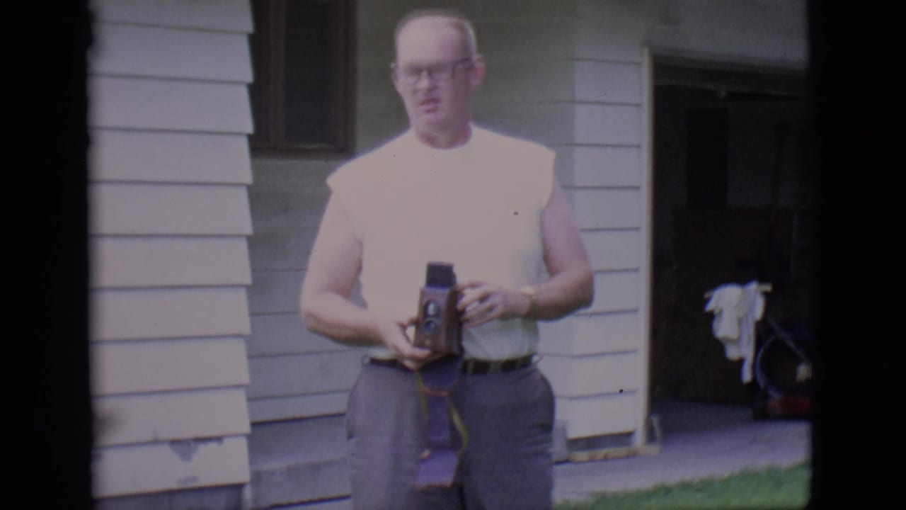 Man stands near house siding and holds a small movie camera at chest height.