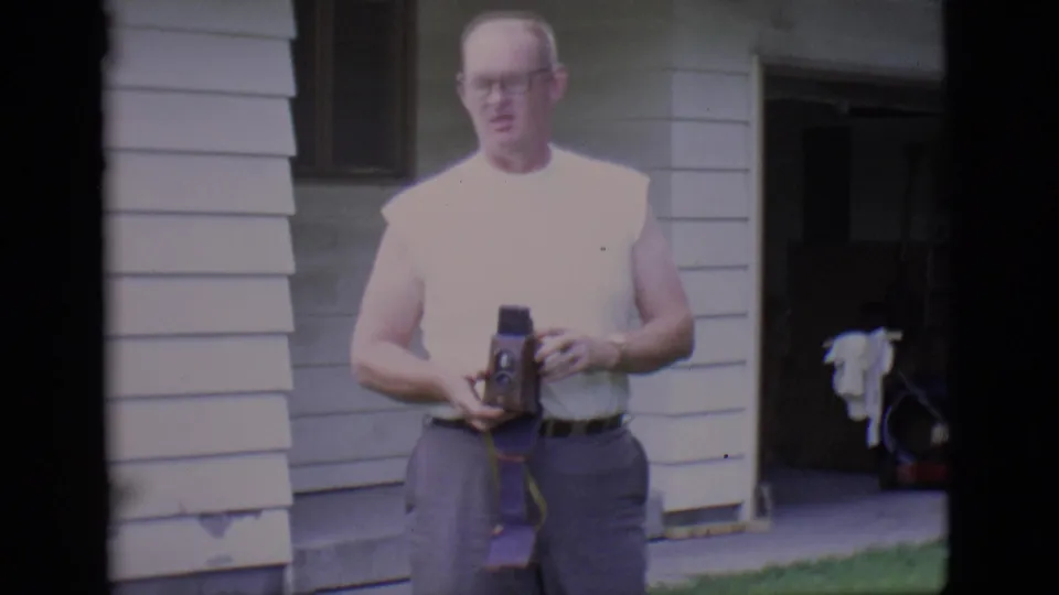 The adult man stands by the Clarkdale house exterior, holding a small movie camera while facing toward the filming position.