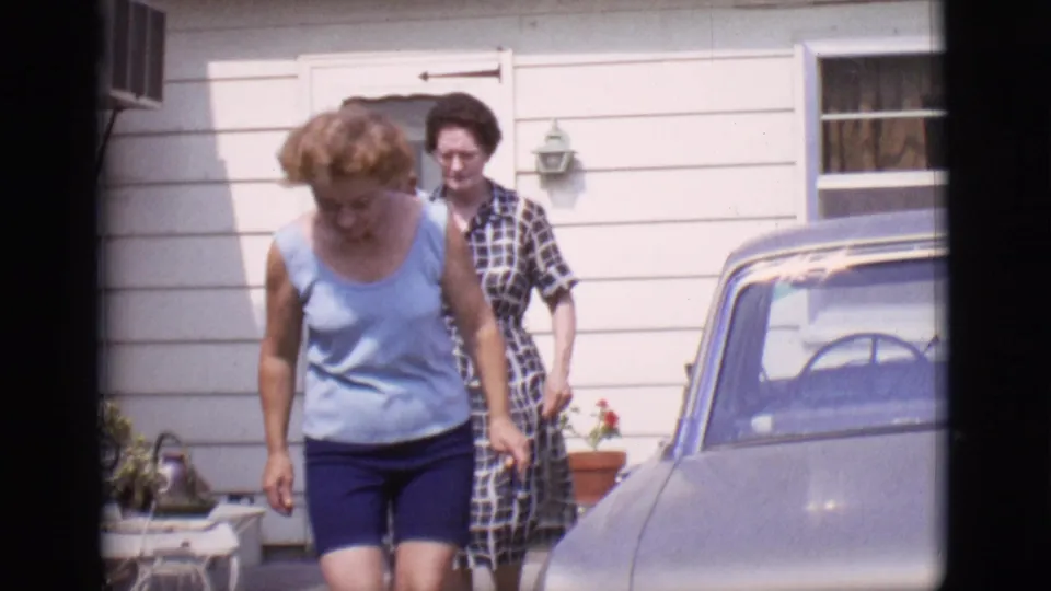 Two adult women walk from a white-sided house toward a parked car in the supplied 1966 Clarkdale, Arizona home-movie scene.