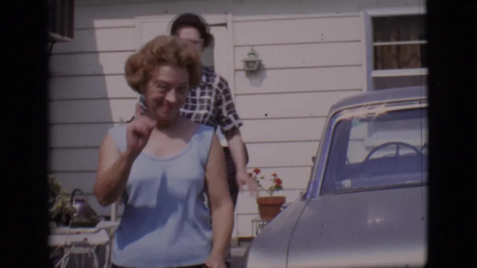 A woman in a light sleeveless top smiles and gestures toward the camera near a driveway car, with another woman behind her in the 1966 home-movie scene.