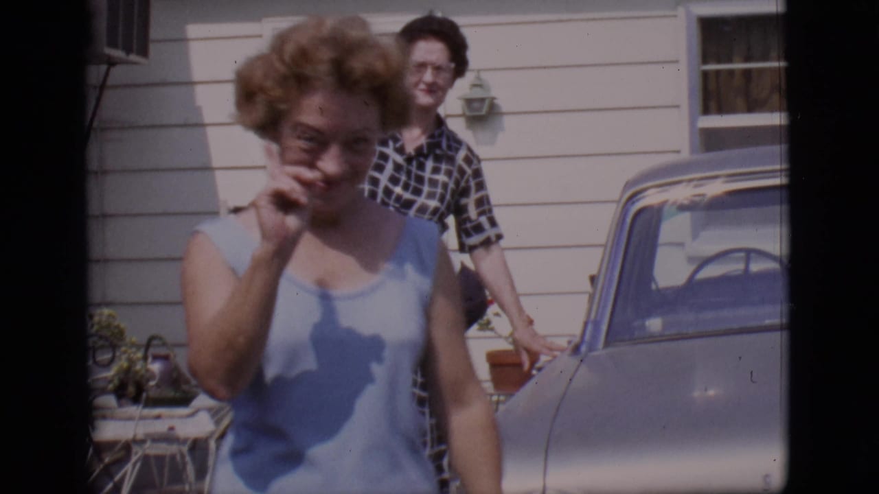 Woman in a light blue top gestures near a parked sedan while another woman stands by the house.