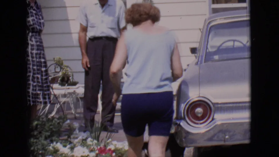 At the end of the clip, the woman walks around the parked sedan with adults, flowers, and house siding in view.