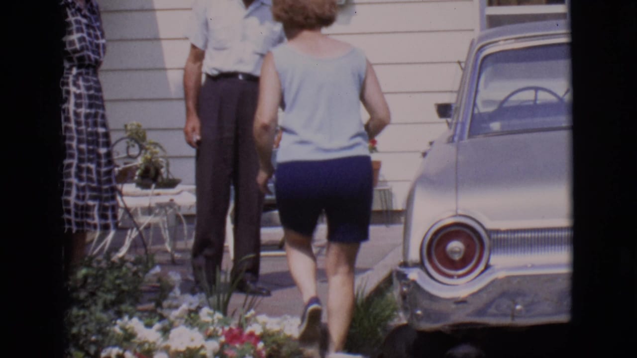 Woman walks toward two adults outside a house beside a parked silver car.