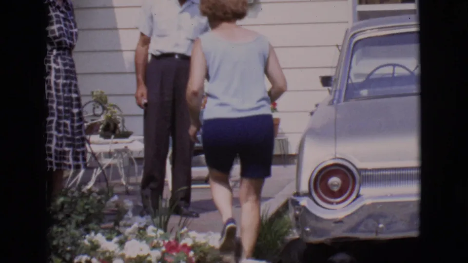 In 1966 Clarkdale, Arizona, a woman in shorts approaches two adults near a white-sided house and parked silver car.