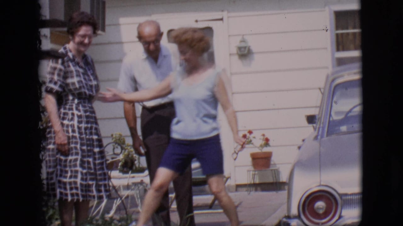 Three adults laugh and greet each other outside a house near a parked car.