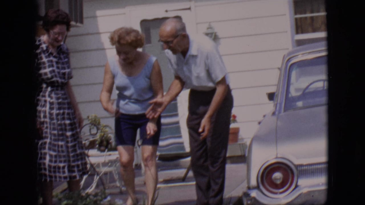Three adults stand near a porch and parked car as they look down toward a small dog.