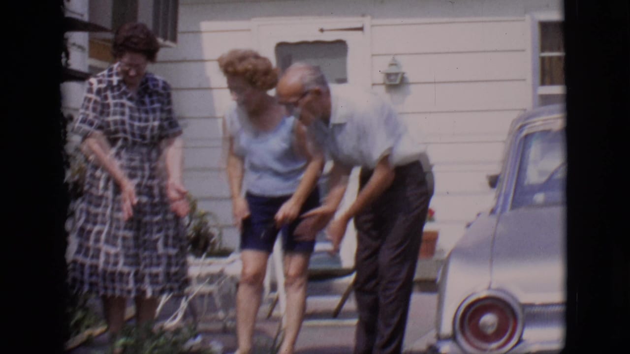Two women and a man lean toward a small dog outside a white-sided house.