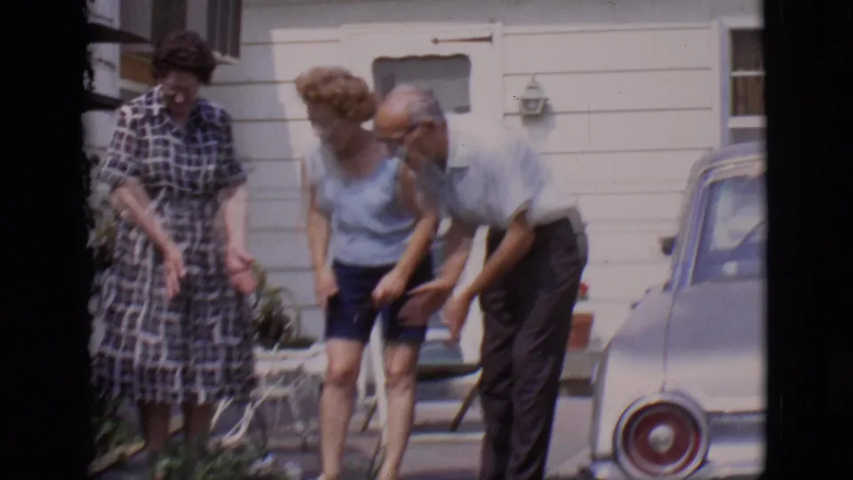Three adults in Clarkdale, Arizona appear focused on a small dog near a porch, with a 1960s car partly visible beside them.