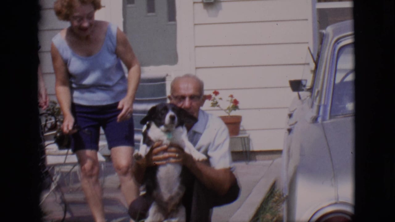 Older man crouches holding a black and white dog while a woman stands nearby outside a house.
