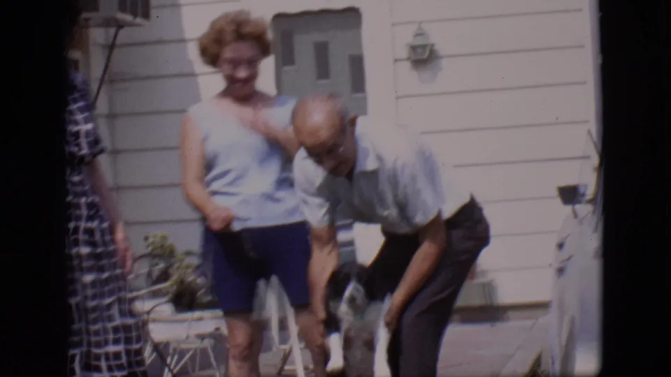 An older man bends down toward the black and white dog as the woman stands close by in the 1966 Clarkdale backyard home-movie scene.