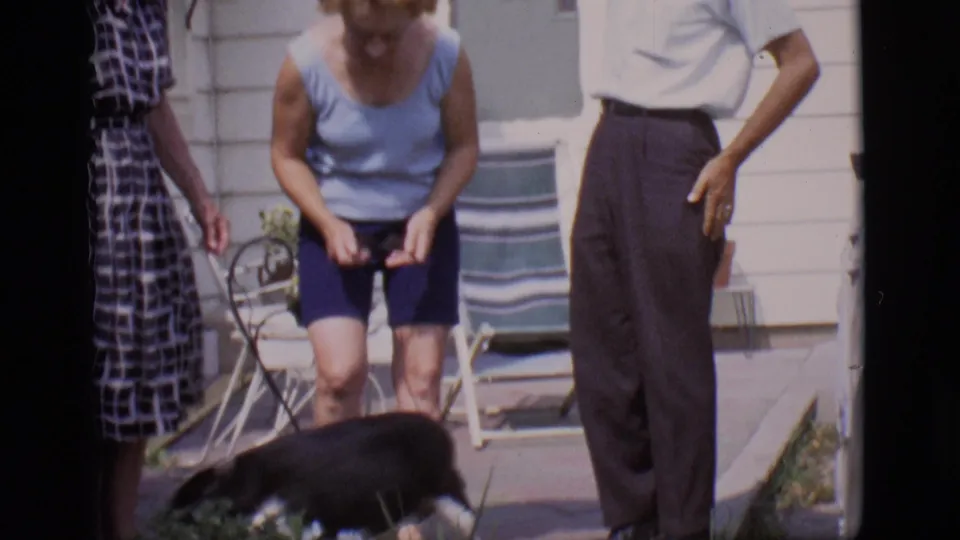 The dog moves across the foreground while adults stand near a lawn chair, doorway, and light house siding in the 1966 Clarkdale home movie.