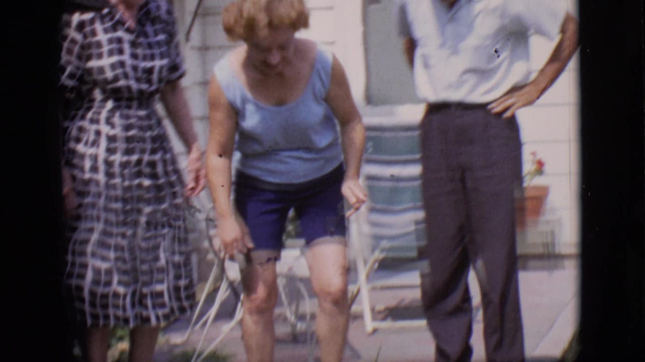 Woman in blue bends toward a small dog while two adults stand near a white house.