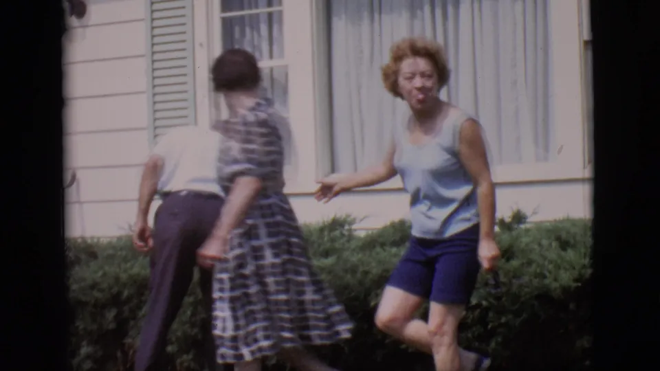 The woman in a blue sleeveless top stands near low bushes while a man and a woman in a checkered dress turn by the house.