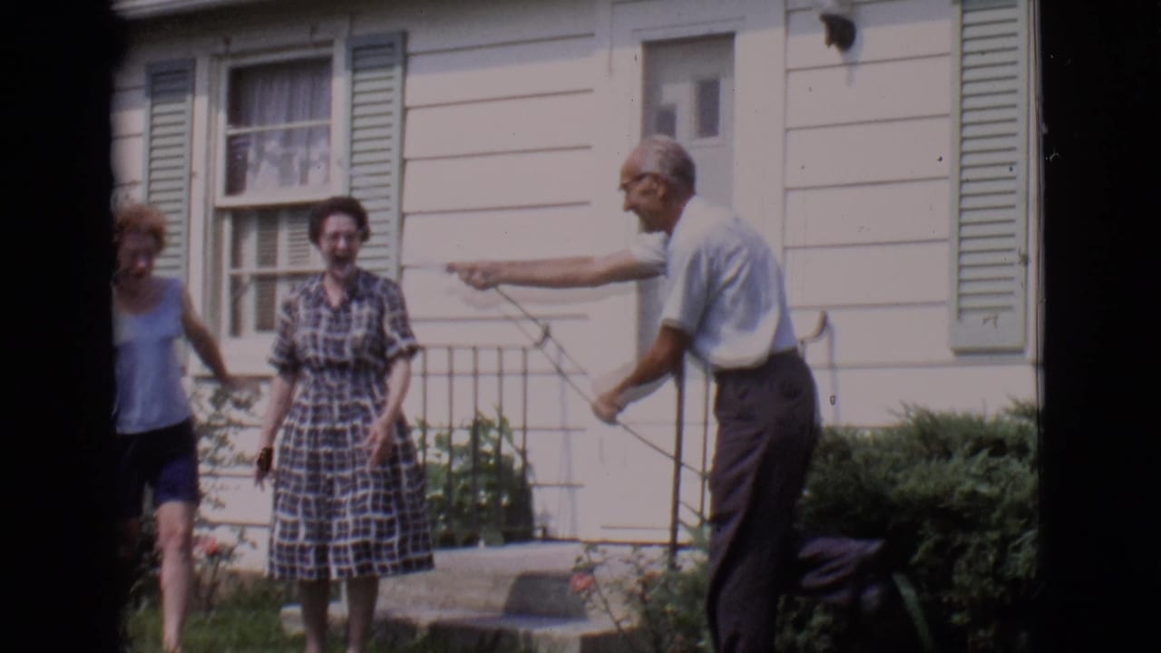Older man points a garden hose toward two women standing by a white house.