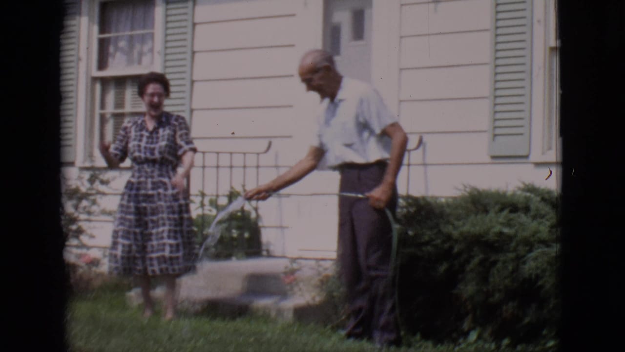 Woman in a plaid dress reacts while an older man sprays a garden hose nearby.