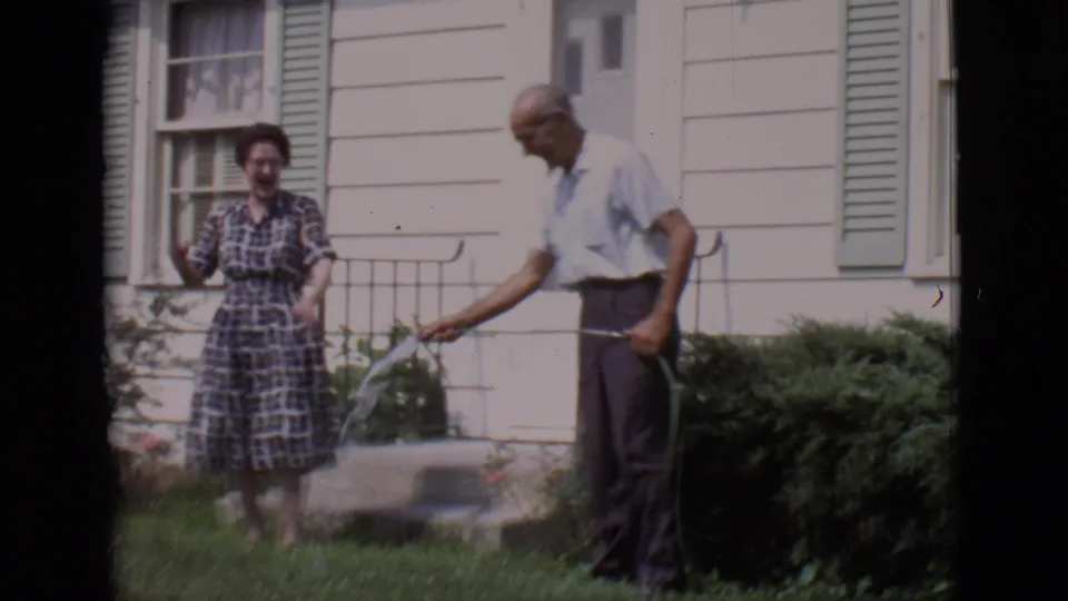 A woman in a plaid dress smiles near the front steps as an older man holds a spraying hose in the 1966 Clarkdale yard scene.