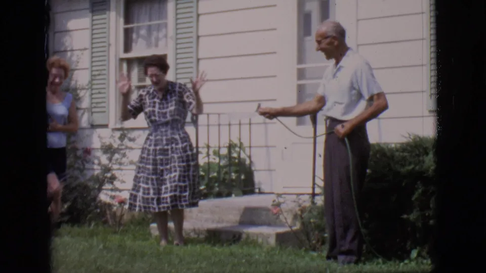Two women react with raised hands while an older man stands with a garden hose outside a white house in Clarkdale, Arizona, 1966.