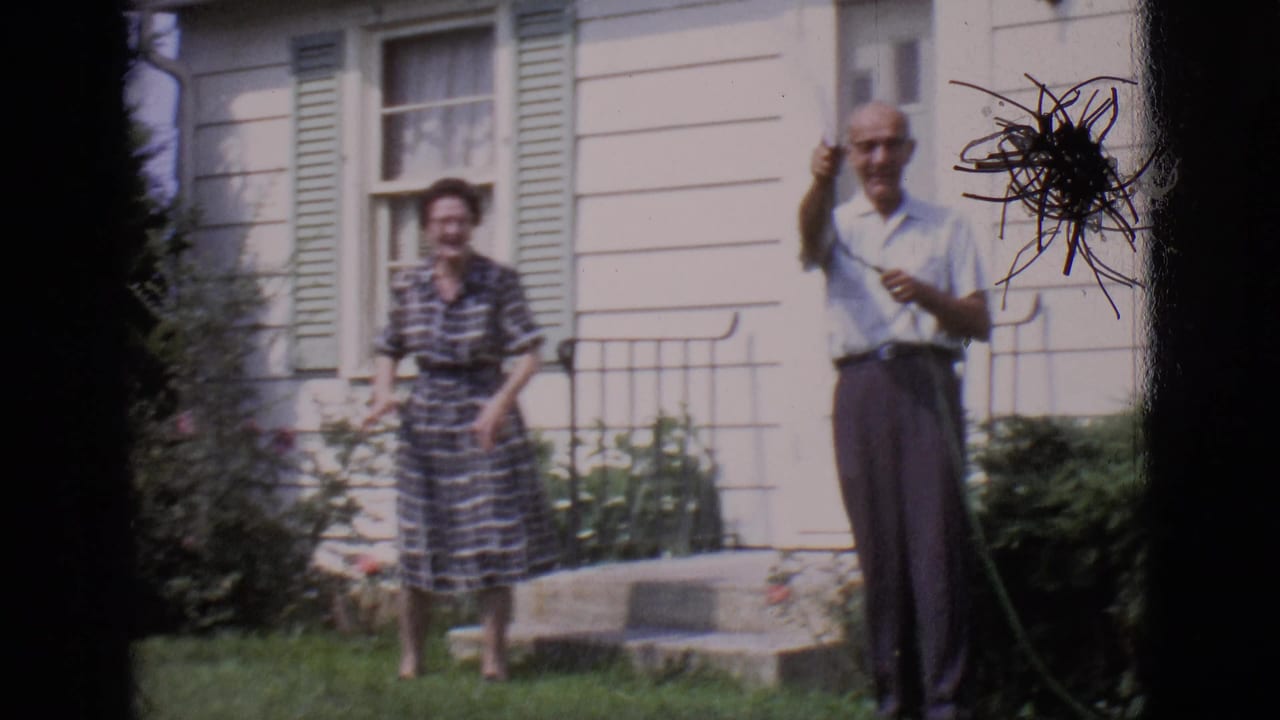 Two adults stand on a lawn by a house while a man holds a garden hose.