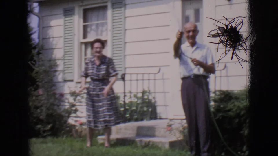 In the 1966 Clarkdale home-movie frame, a woman in a plaid dress and a man with a hose stand outside a pale house.