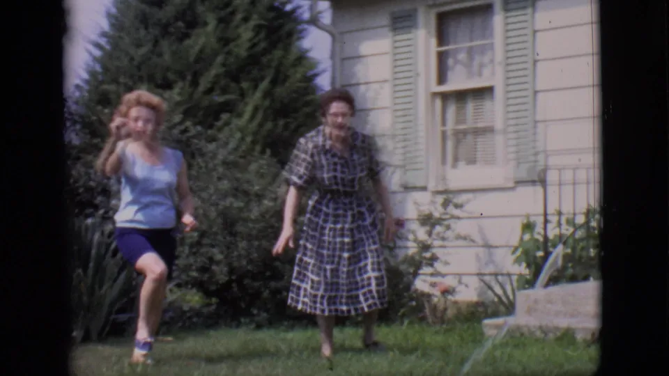 In the Clarkdale 1966 yard scene, a woman in a sleeveless top approaches as a woman in a plaid dress stands near the house.