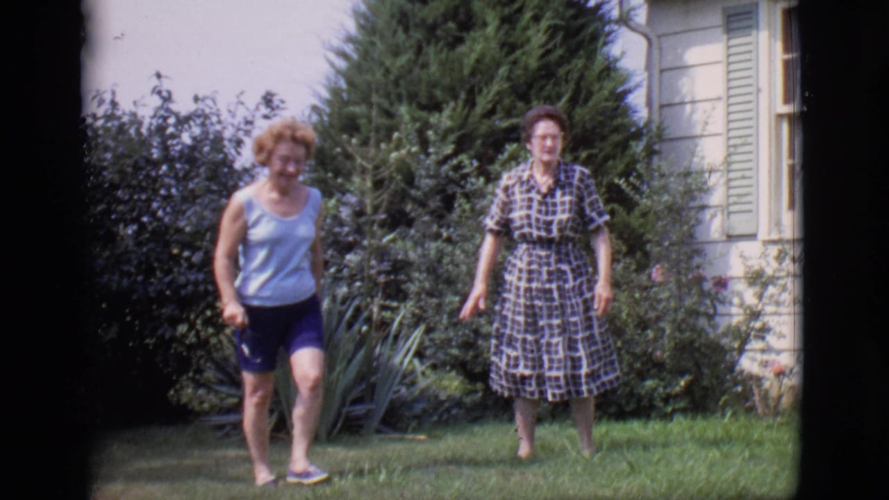 Two women walk across a backyard lawn near shrubs and a white house with green shutters.