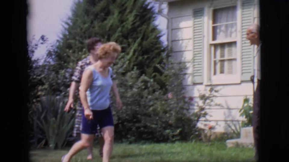 A woman in a light sleeveless top walks across the Clarkdale backyard while another woman is partly obscured near the house and shrubs.